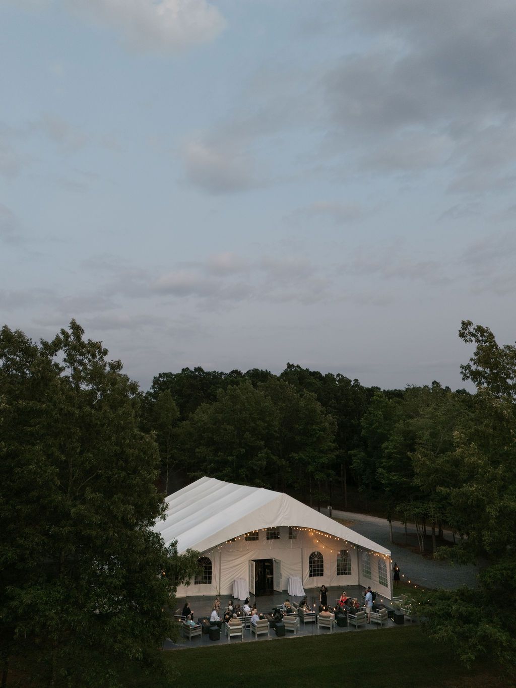 White wedding tent pavilion with guests seated for outdoor reception at dusk surrounded by green forest trees.