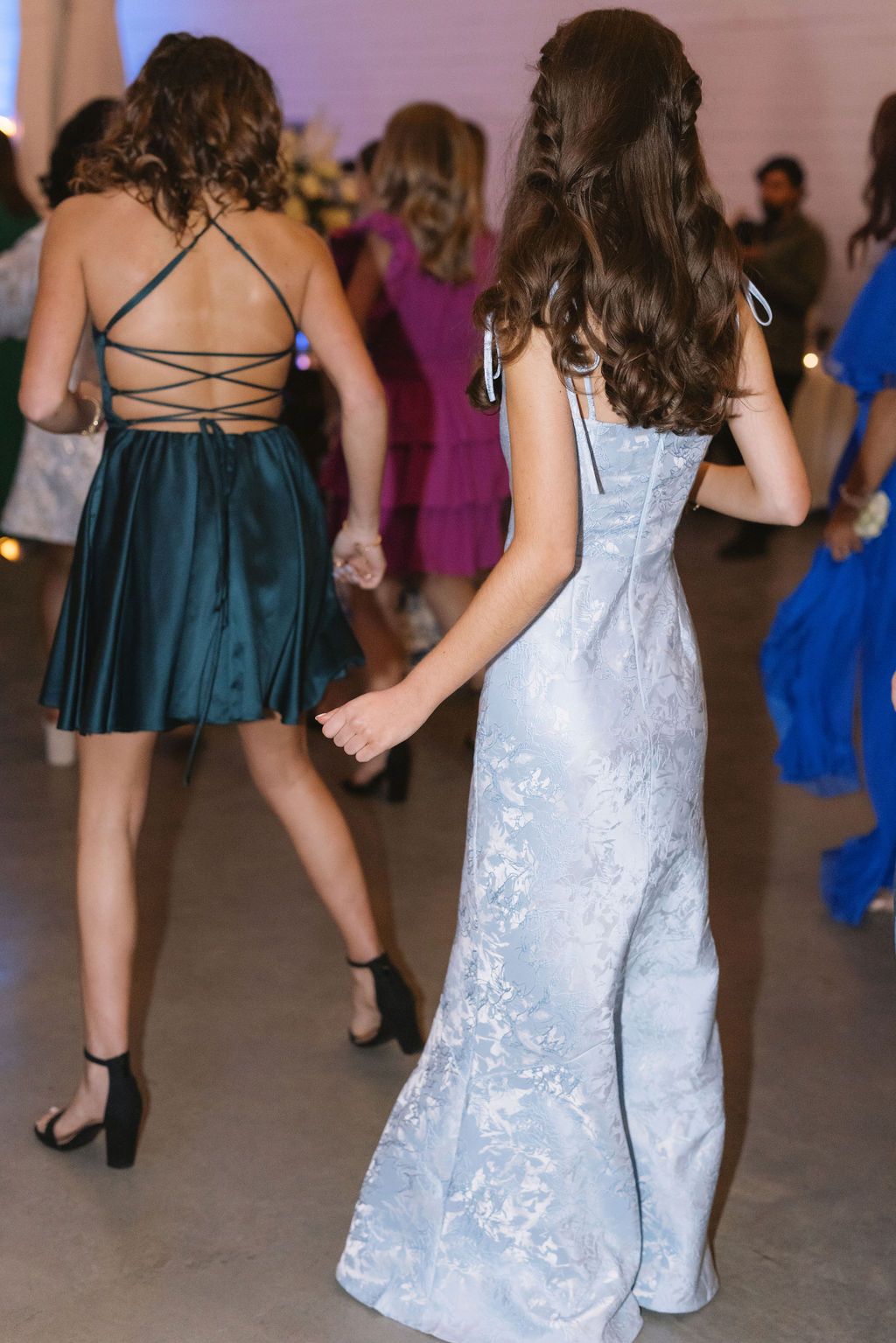 Partygoers in formal attire dancing at an event, featuring a long white floral dress and a teal strappy cocktail dress.