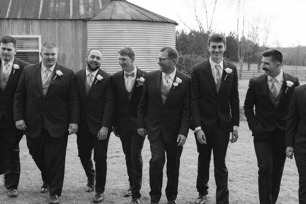 A group of groomsmen in dark suits walk together in a black and white photo during a wedding celebration.