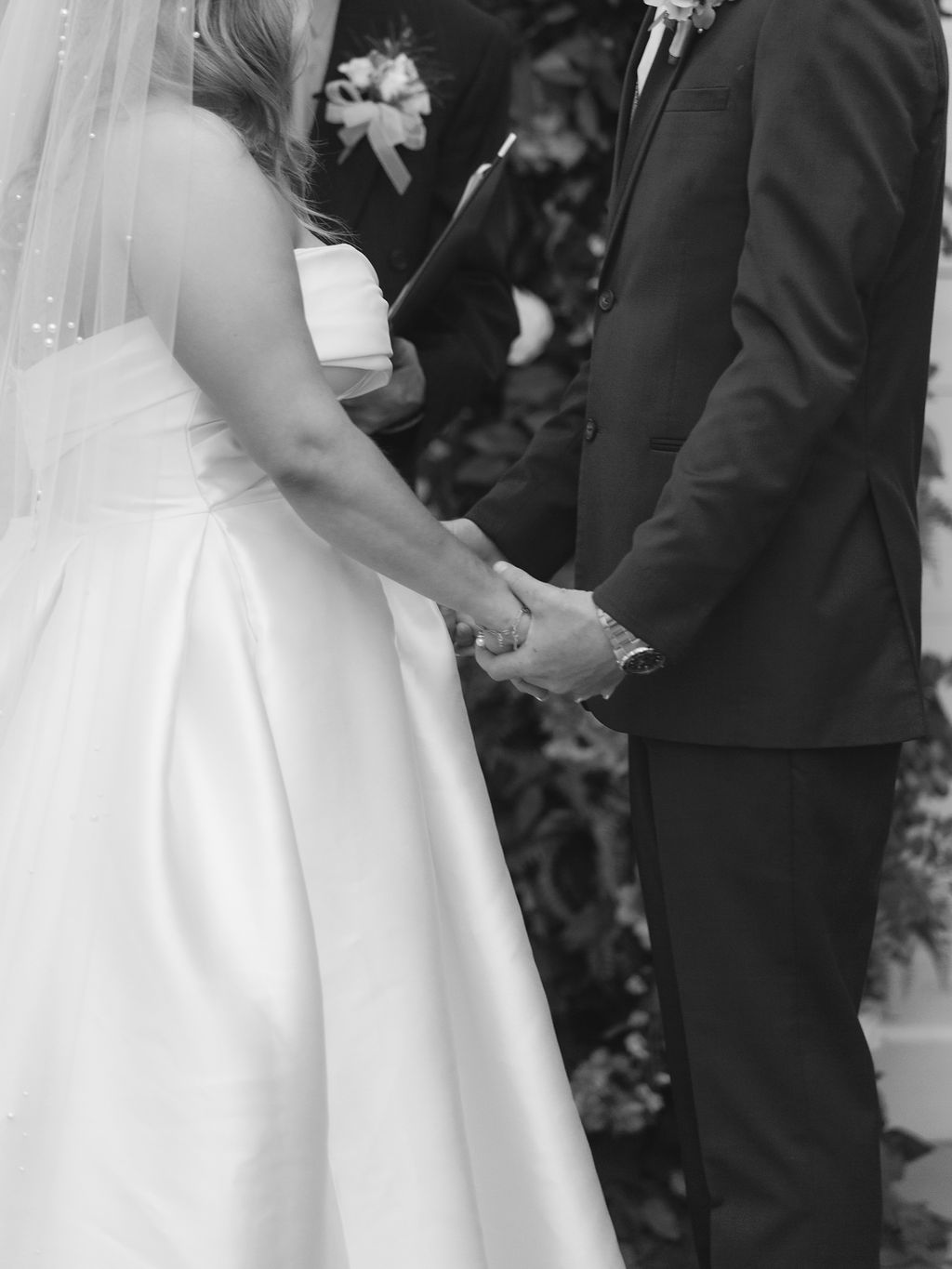 A black and white wedding ceremony photo showing a couple holding hands during their vows in formal attire.