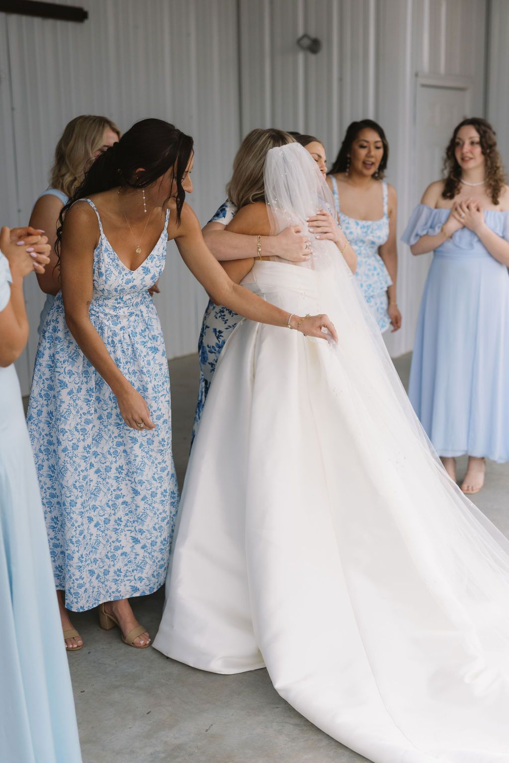 Bridesmaids in light blue floral dresses gather around bride in white wedding gown during pre-ceremony moments.