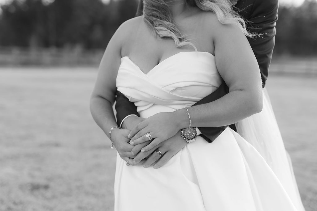 Black and white photo showing couple embracing, with focus on wedding dress and clasped hands.