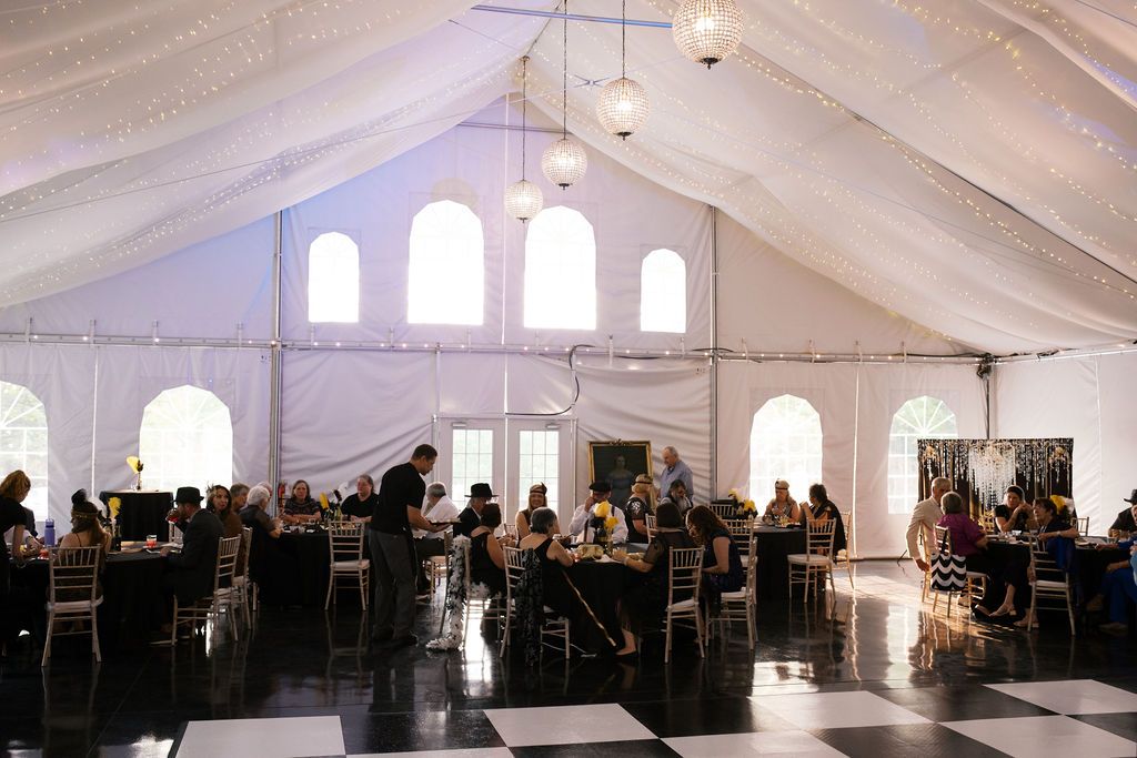 An elegant white tent reception venue with cathedral windows, black and white checkered floor, and round dining tables.