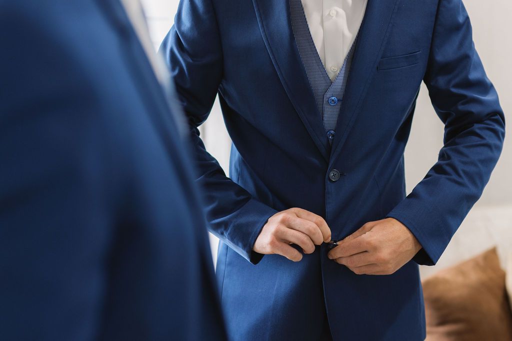 A person wearing a navy blue suit adjusts their jacket button while standing.