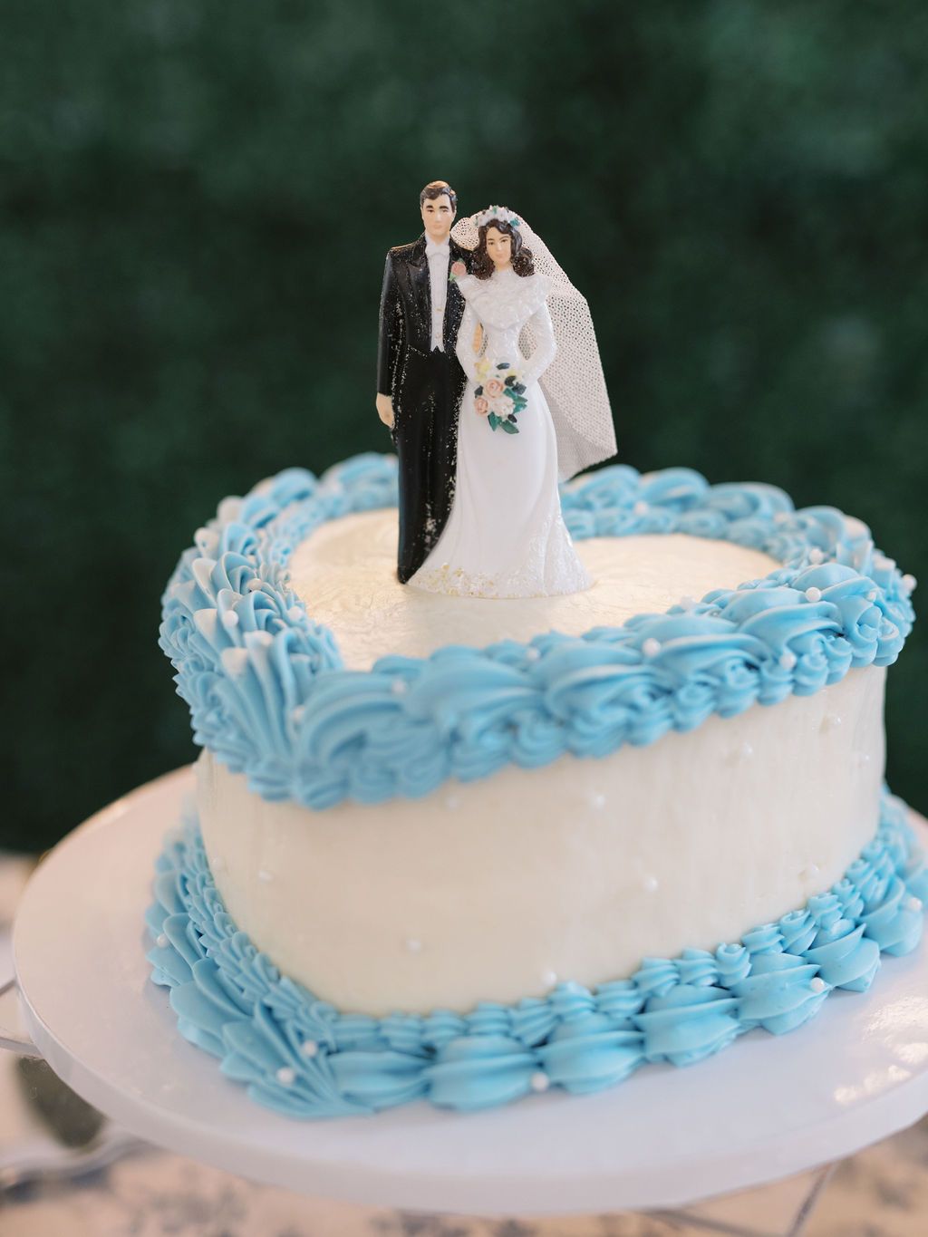 A white wedding cake decorated with blue piped frosting trim and a traditional bride and groom cake topper.