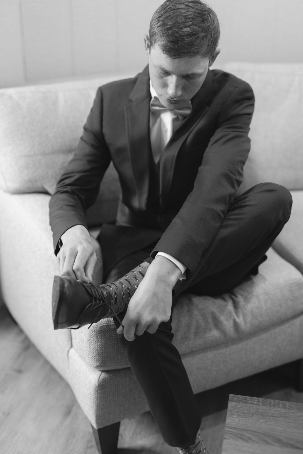 A businessman in a black suit sits on a couch while adjusting his dress boots in this black and white photo.