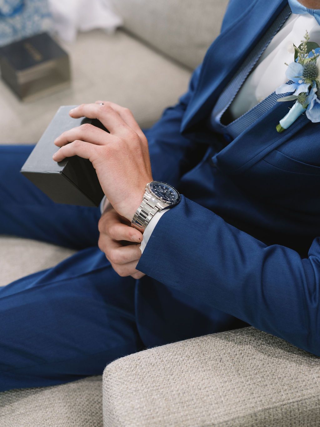 Close-up of a person in a navy blue suit wearing a silver watch and boutonniere while seated on a grey couch.