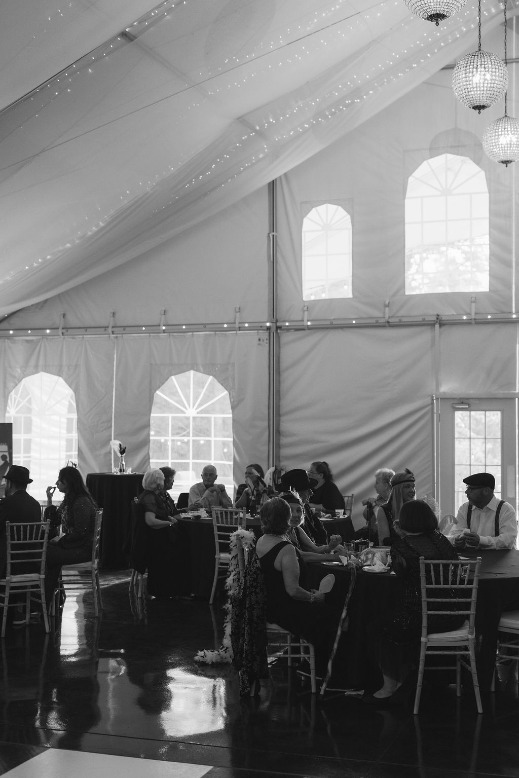 Black and white photo of wedding reception guests dining under an elegant tent with cathedral windows and string lights.