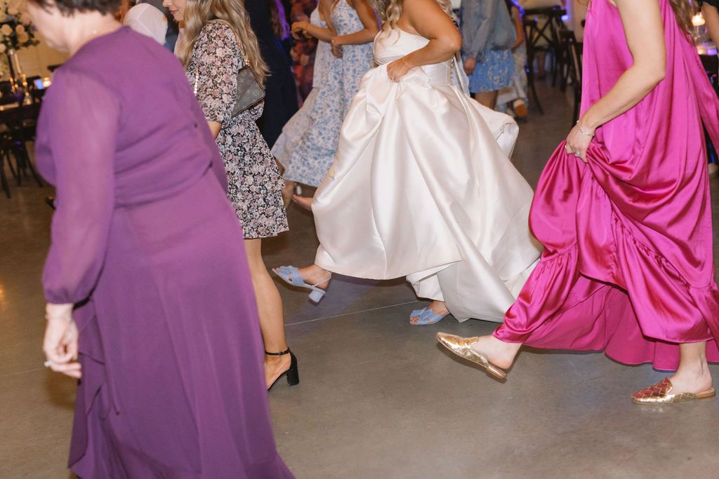People dancing at a wedding reception wearing colorful formal dresses in purple, white and pink.