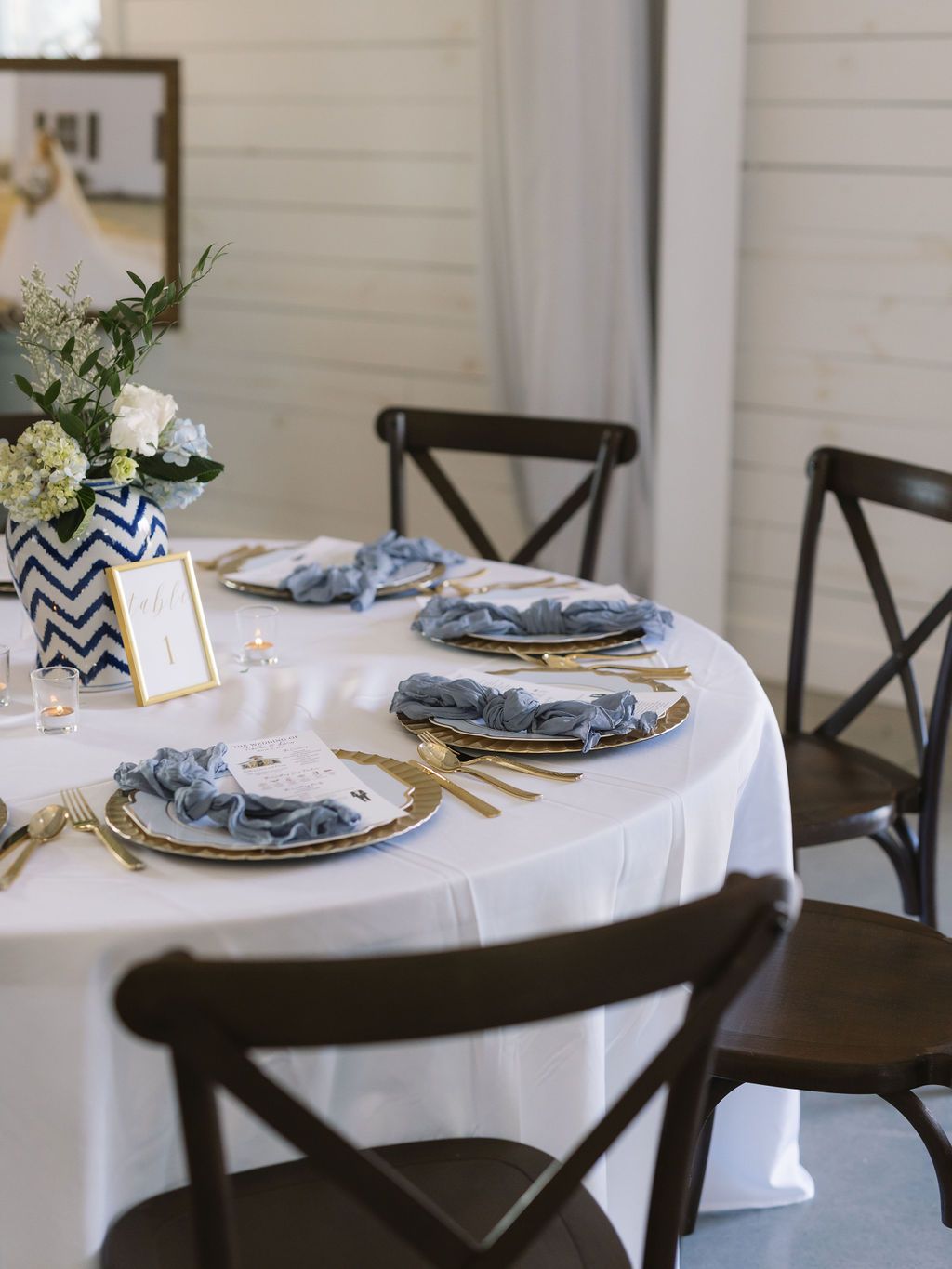 Elegant table setting with white tablecloth, dark wooden chairs, gold chargers, and blue napkins for intimate dining.