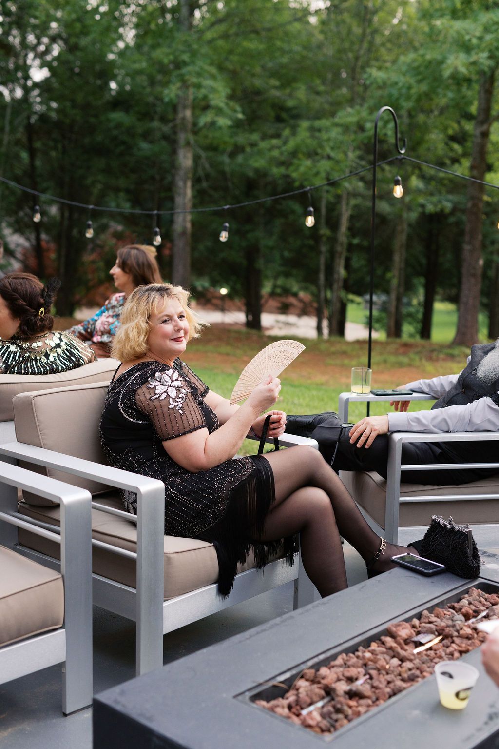 Guests relax on modern outdoor furniture with string lights overhead and wooded scenery in the background.