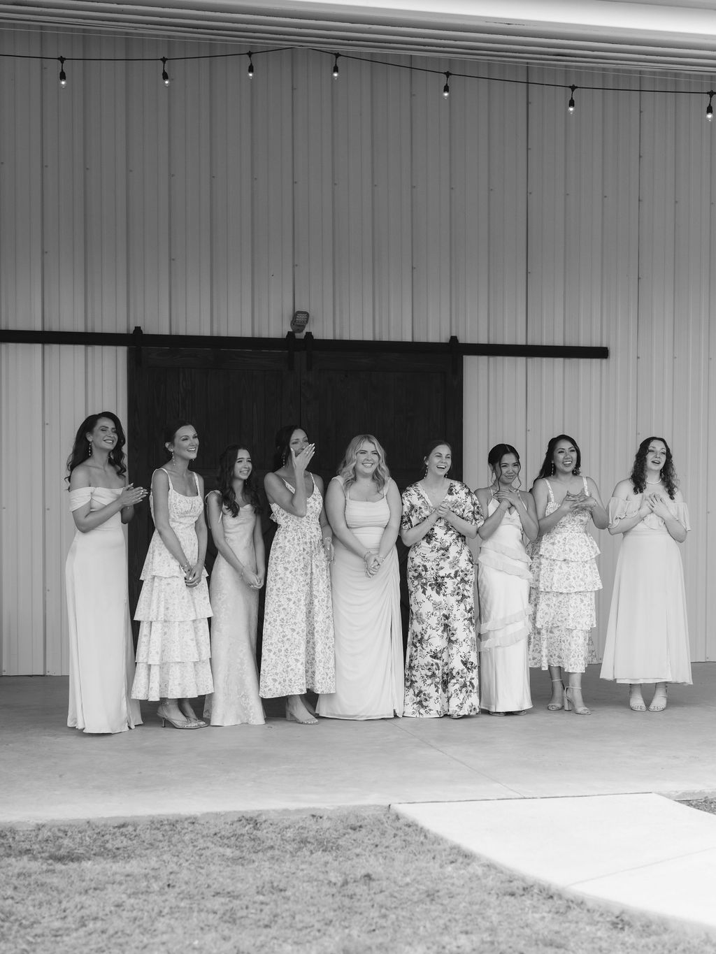 A group in light-colored dresses stands in a line against a paneled wall with a sliding barn door.