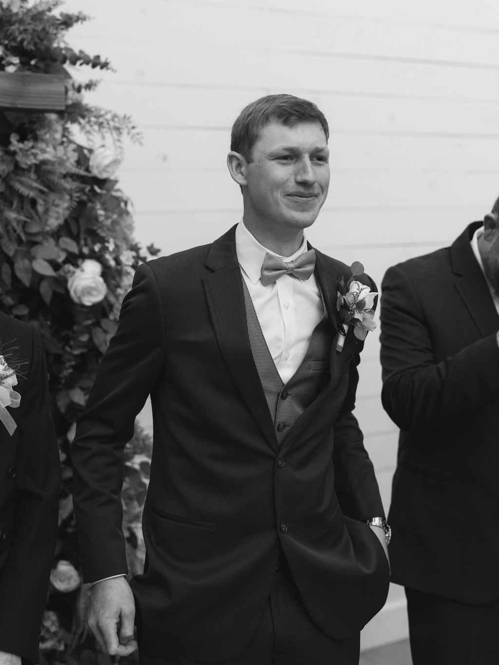 A groom in a classic black tuxedo and bowtie stands at a wedding ceremony with floral decorations in the background.
