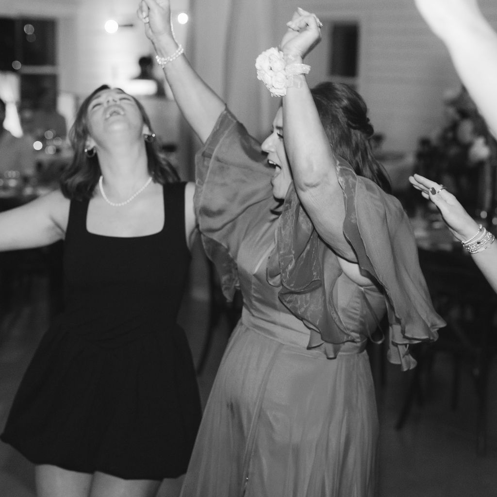 Two women dancing joyfully together at a party in black and white, one in a black dress and one in a flowing gown.