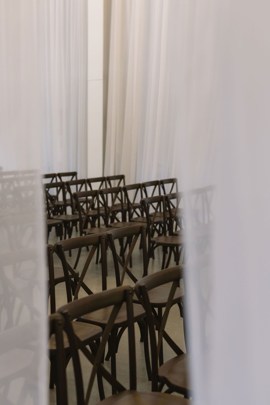 Rows of empty wooden cross-back chairs arranged in a room with white curtains creating a shadowy ambiance.