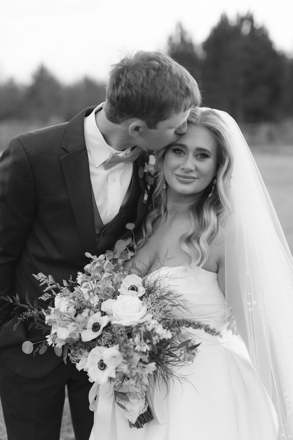 A bride in a white wedding dress holds an elegant bouquet while sharing an intimate moment with the groom outdoors.