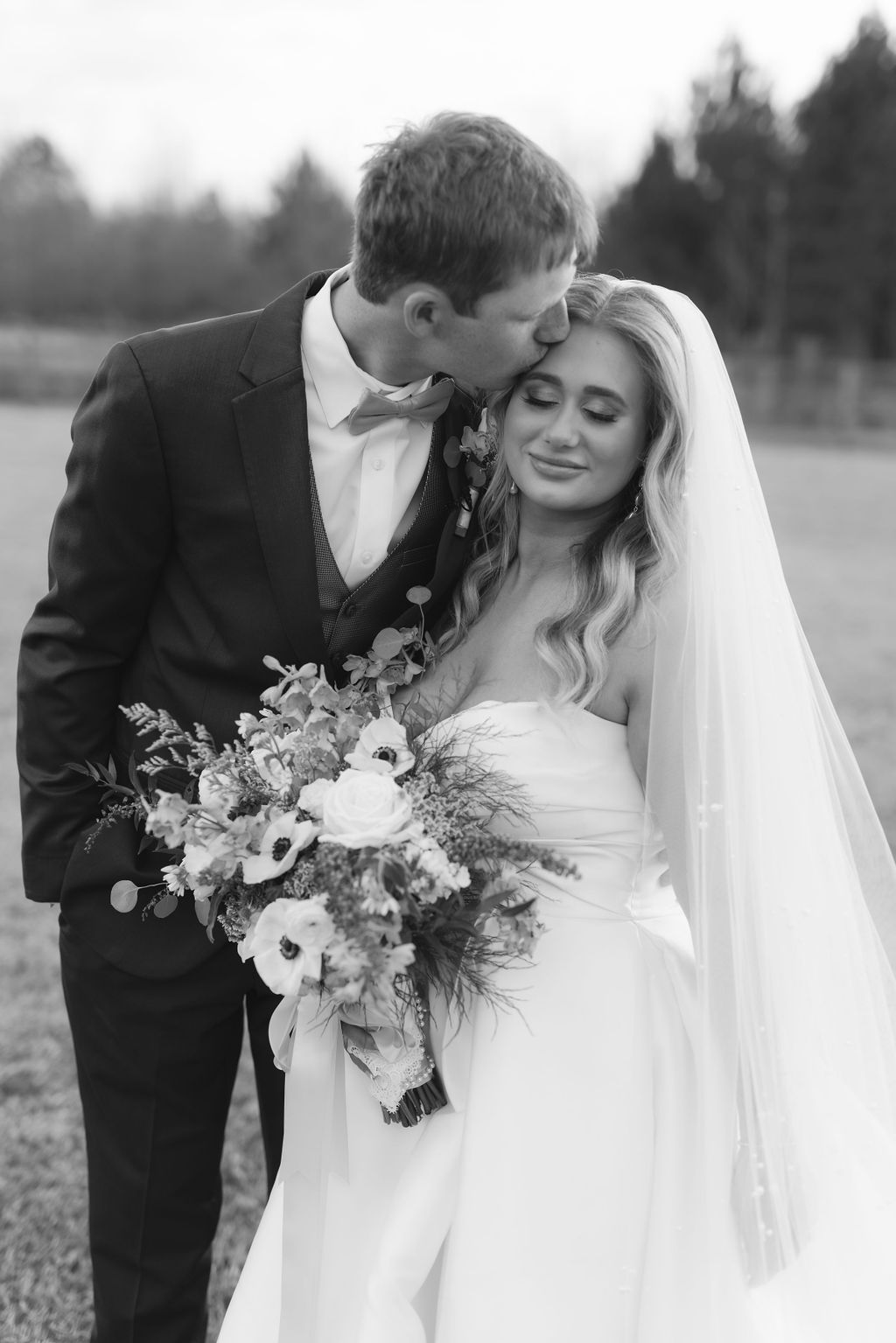 A tender black and white wedding photograph showing a couple sharing an intimate moment with a beautiful floral bouquet.