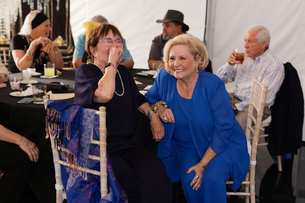 Two people in blue and black outfits share an animated moment of laughter at an indoor social gathering.