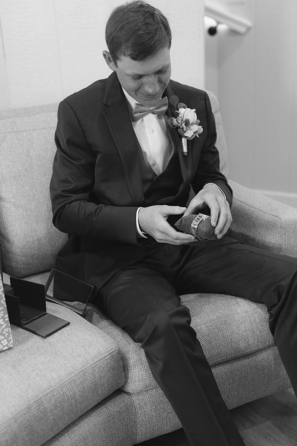 A groom in a black tuxedo and bow tie sits on a couch looking at something in his hands before the wedding ceremony.