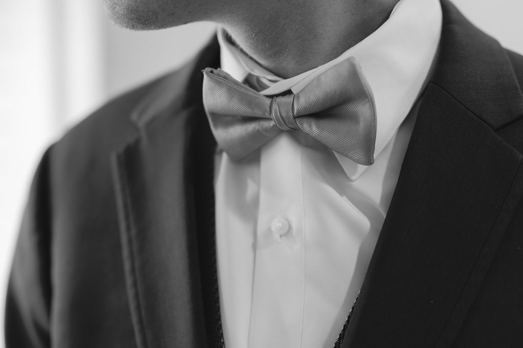 Close-up black and white photo of a gray bow tie worn with crisp white dress shirt and dark suit jacket.