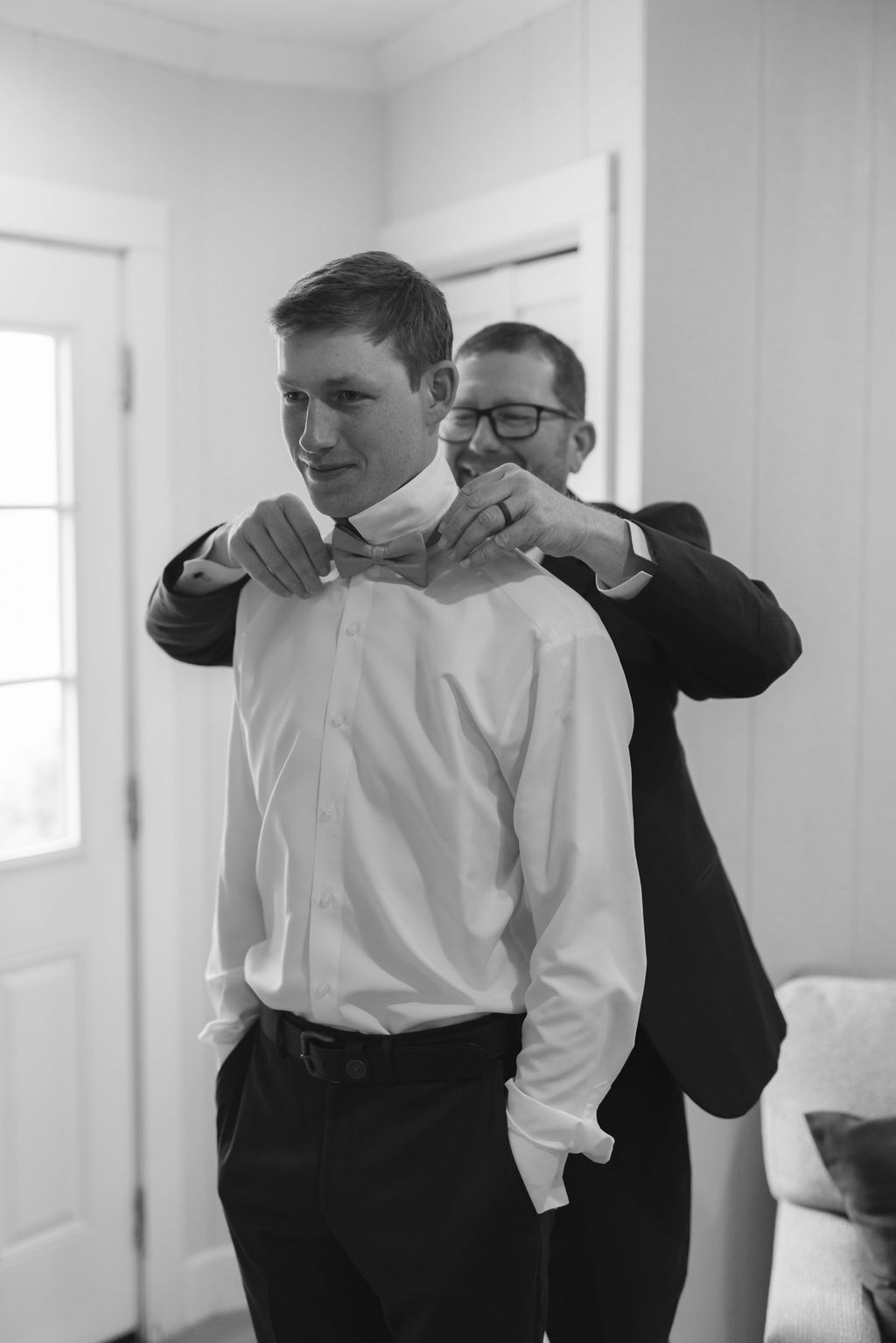 A black and white photo of someone helping adjust a bowtie on another person wearing a white dress shirt.