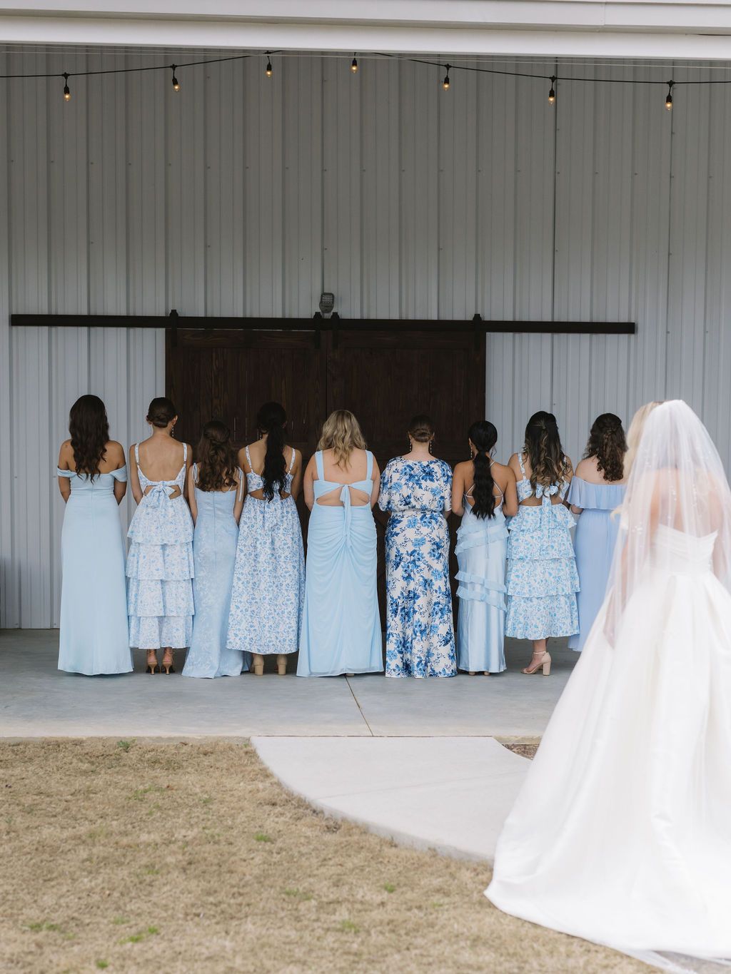 A group of bridesmaids in light blue dresses face a rustic barn door while a bride in white stands to the side.
