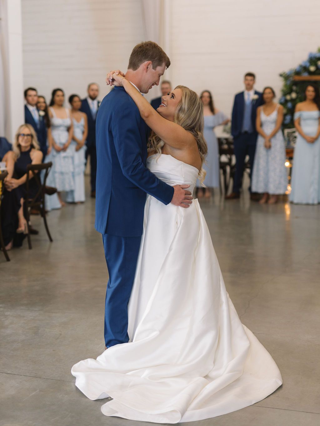A couple shares their first dance as newlyweds in a modern venue with bridesmaids in light blue dresses watching.