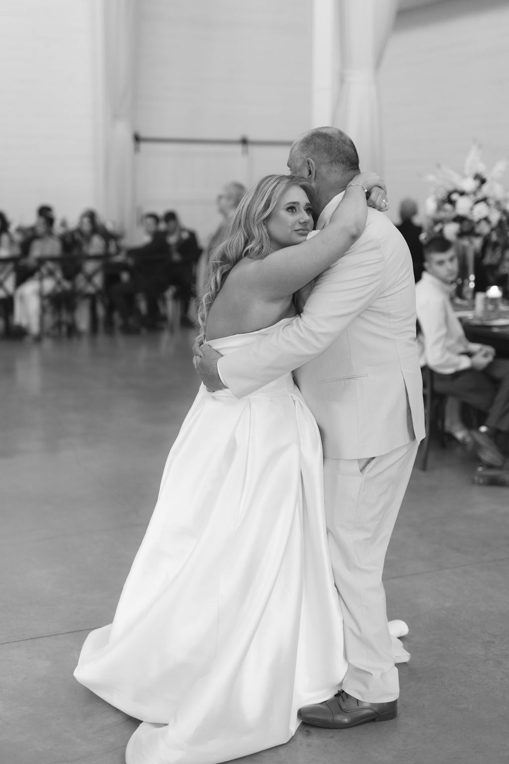 A couple in white attire share their first dance at a wedding reception as guests look on in black and white photo.