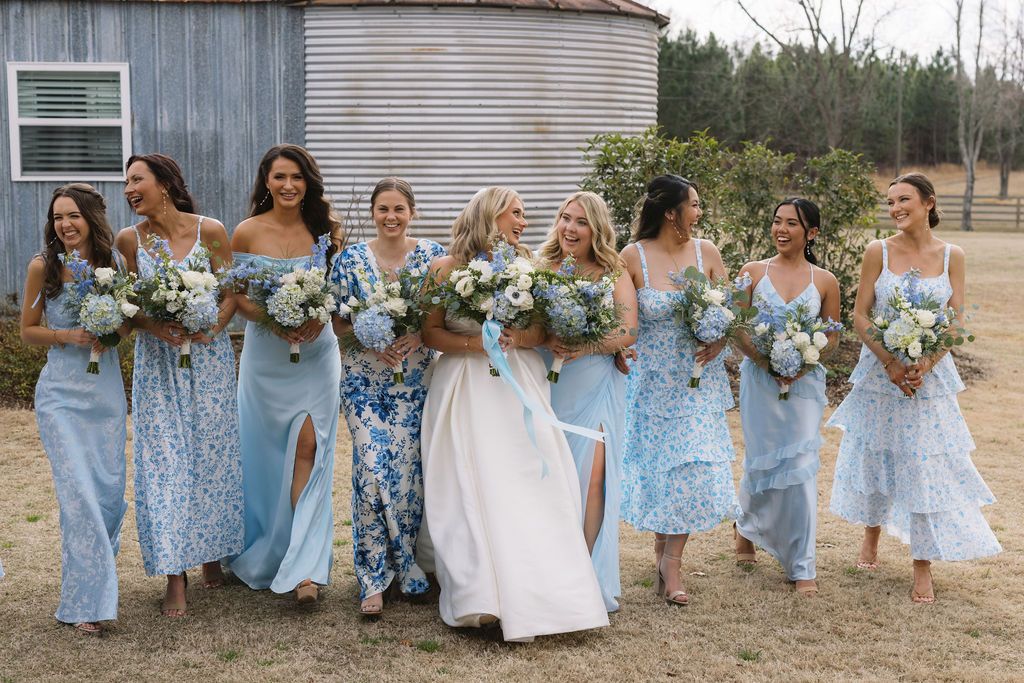 Bridal party in coordinating light blue dresses and gowns walk outdoors while holding floral bouquets near a rustic barn.