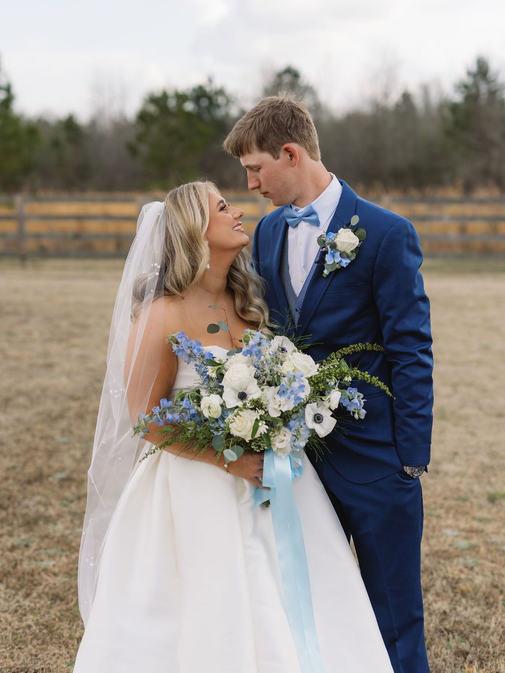 Newlywed couple poses together in a field with the bride holding a blue and white floral bouquet.