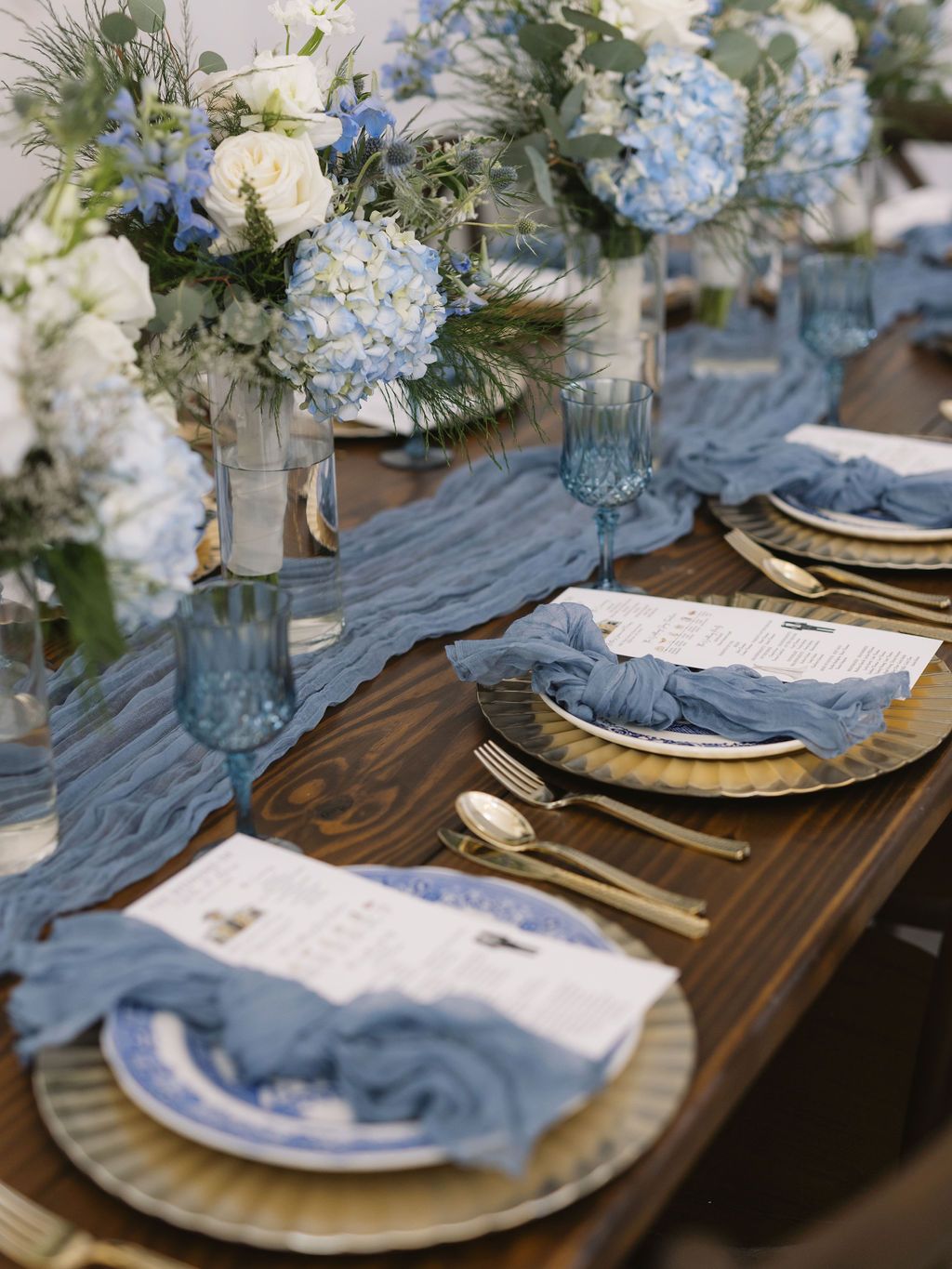 Elegant wedding table setting with blue hydrangeas, gold chargers, and silk runner on rustic wooden table.