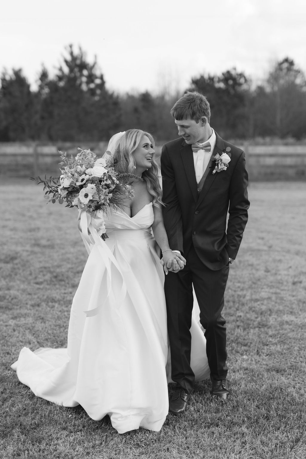Black and white wedding photo of newlyweds in elegant attire holding hands in an outdoor field with trees in background.