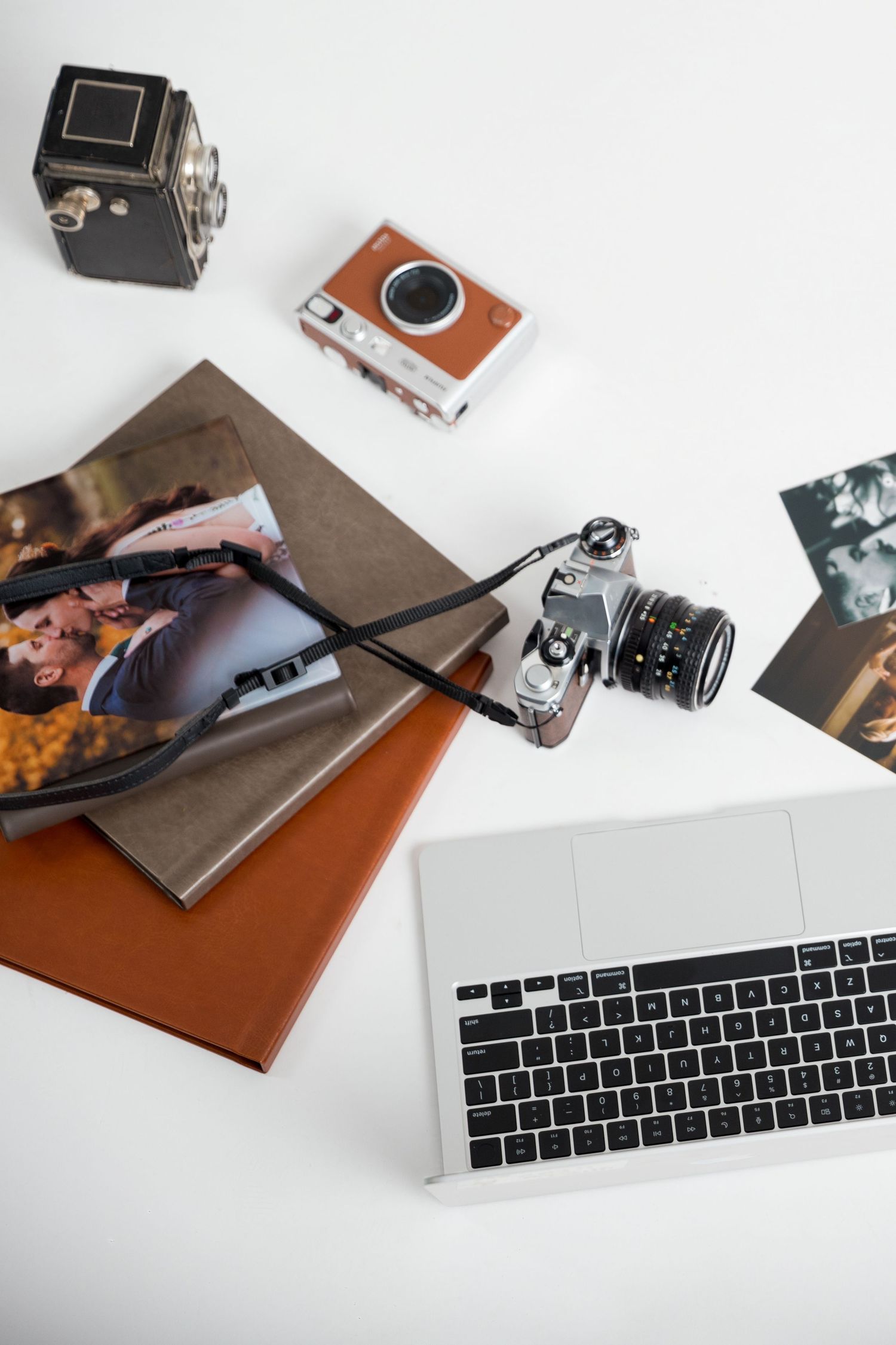 Vintage and modern cameras laid out on white surface next to laptop keyboard and photographs.