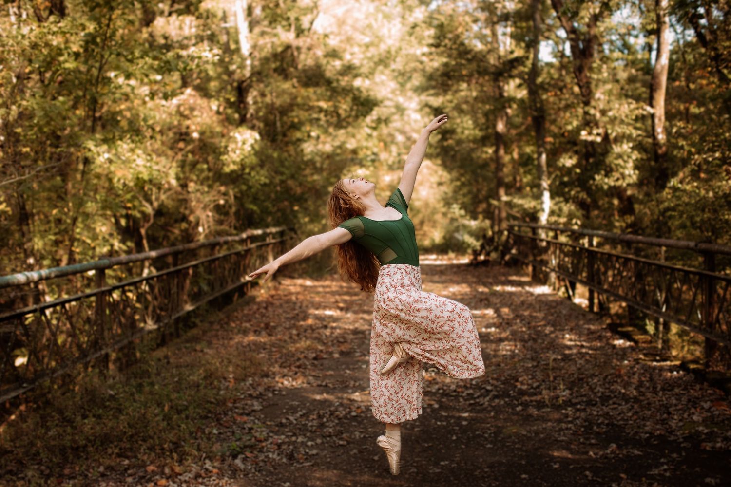 A dancer in a floral dress performs an elegant pose on a rustic wooden bridge surrounded by autumn trees.
