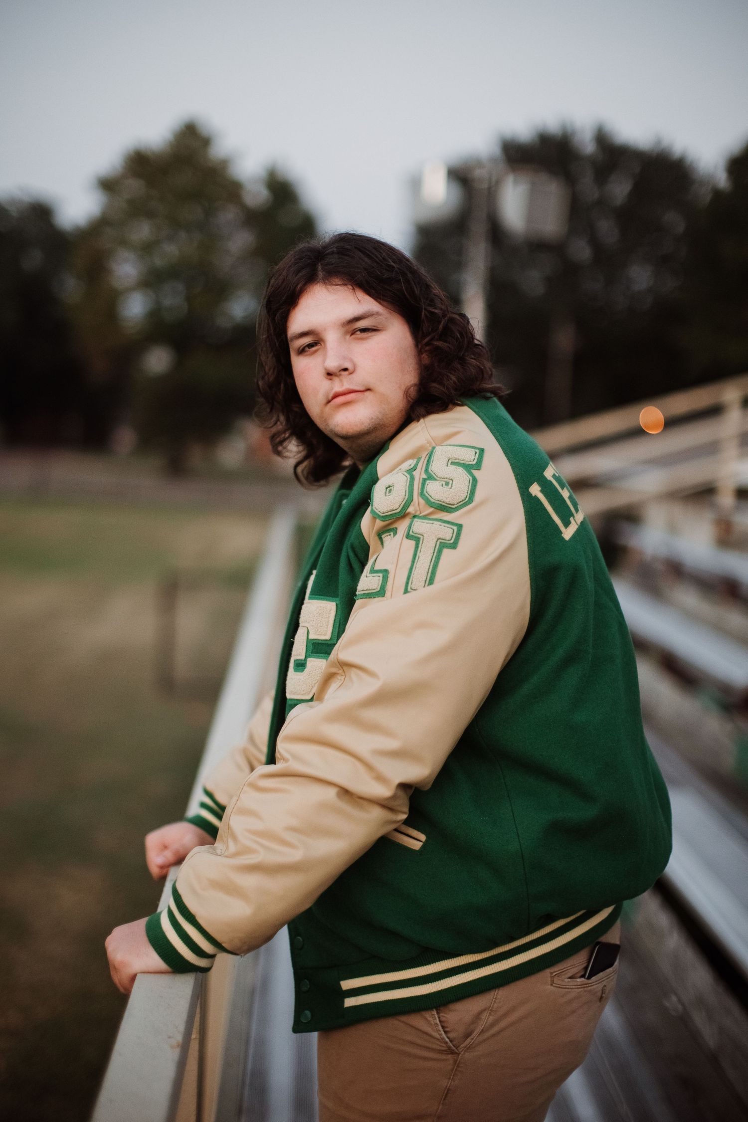 A person wearing a green and cream varsity jacket leans against metal bleacher seating at a sports field.