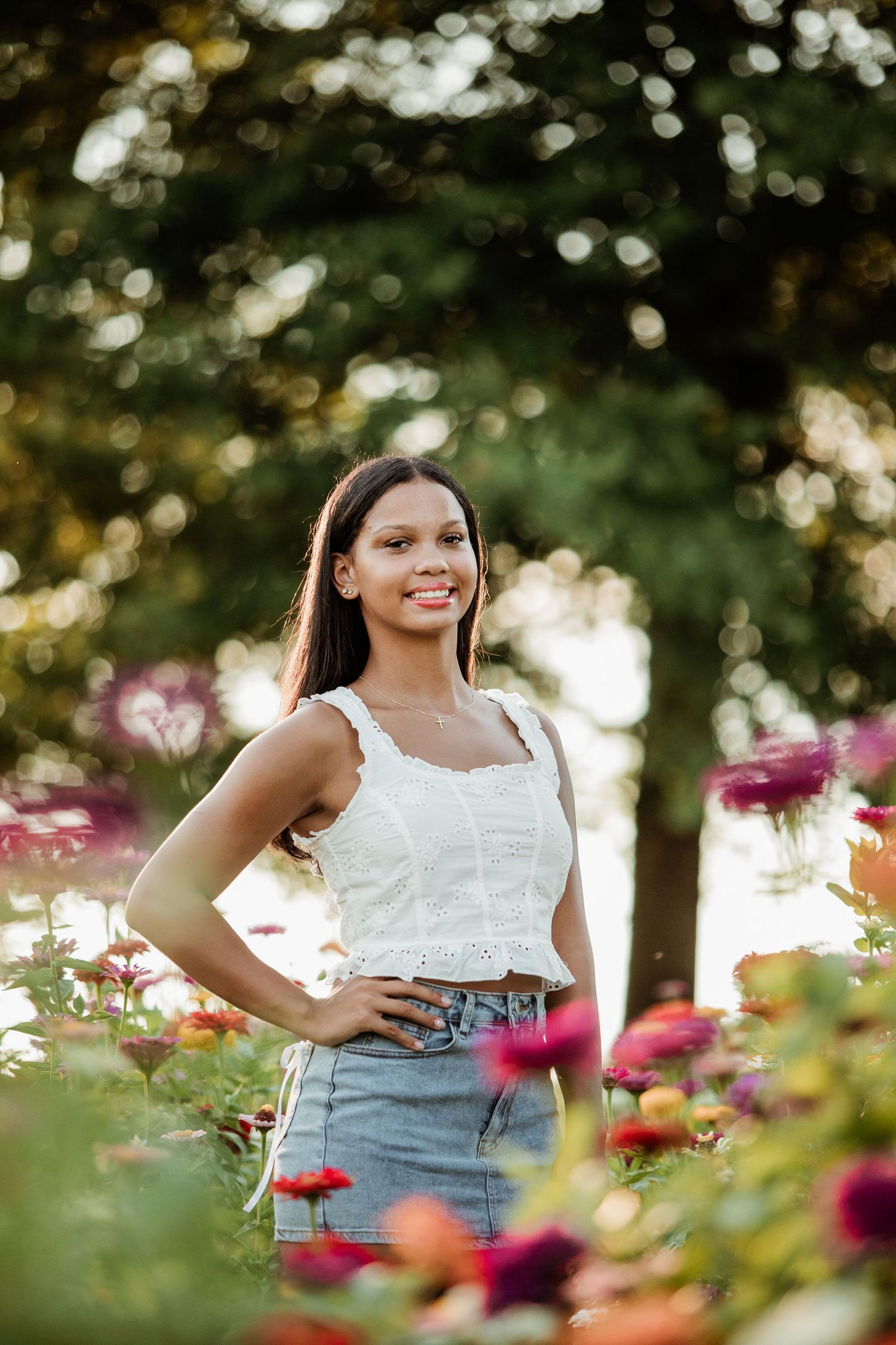 A person poses in a white lacy top and denim skirt among blooming pink flowers in a garden with bokeh lighting effects.