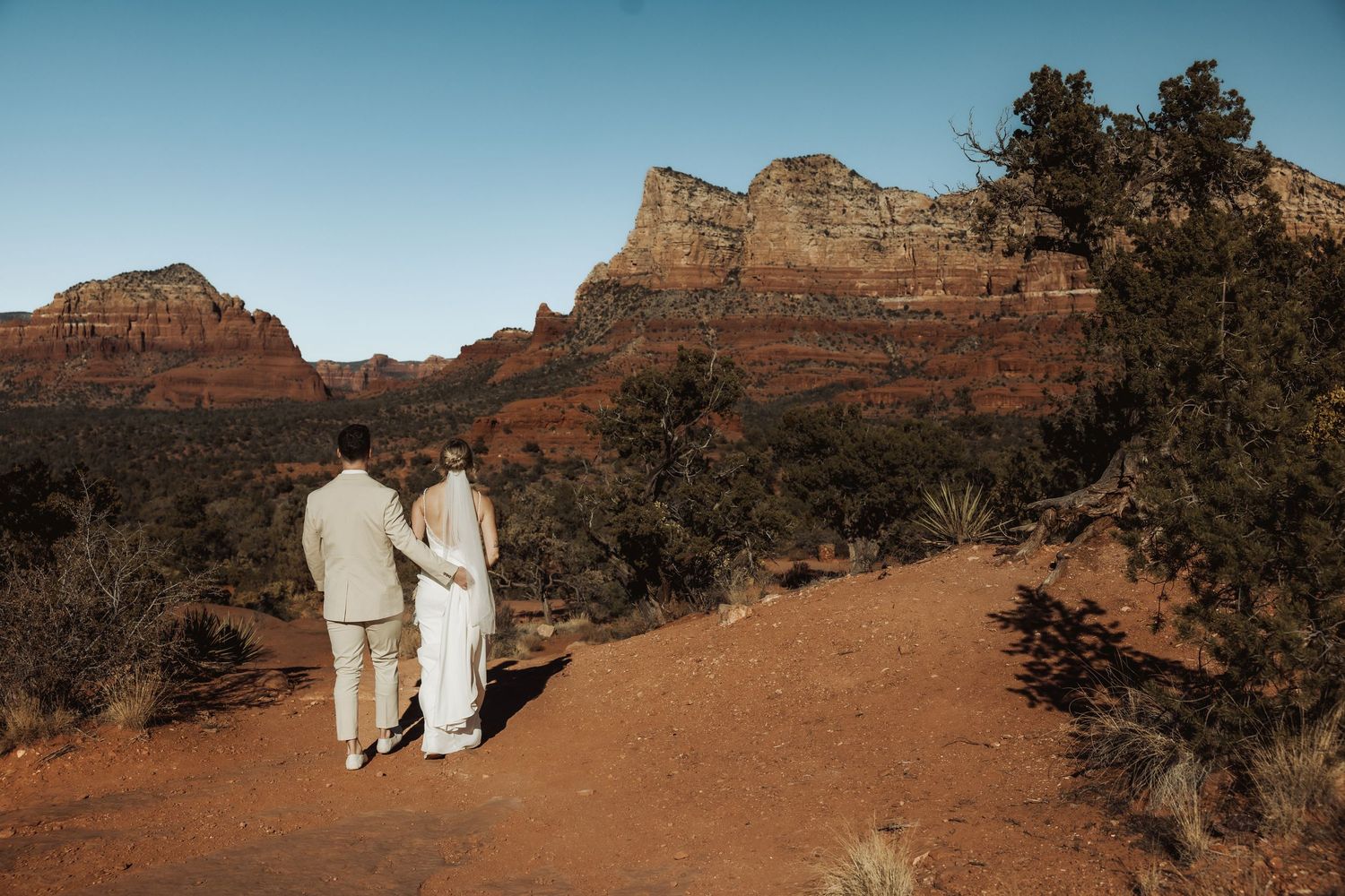 Couple in white attire walking hand in hand on red rock trail with dramatic desert cliffs in background.