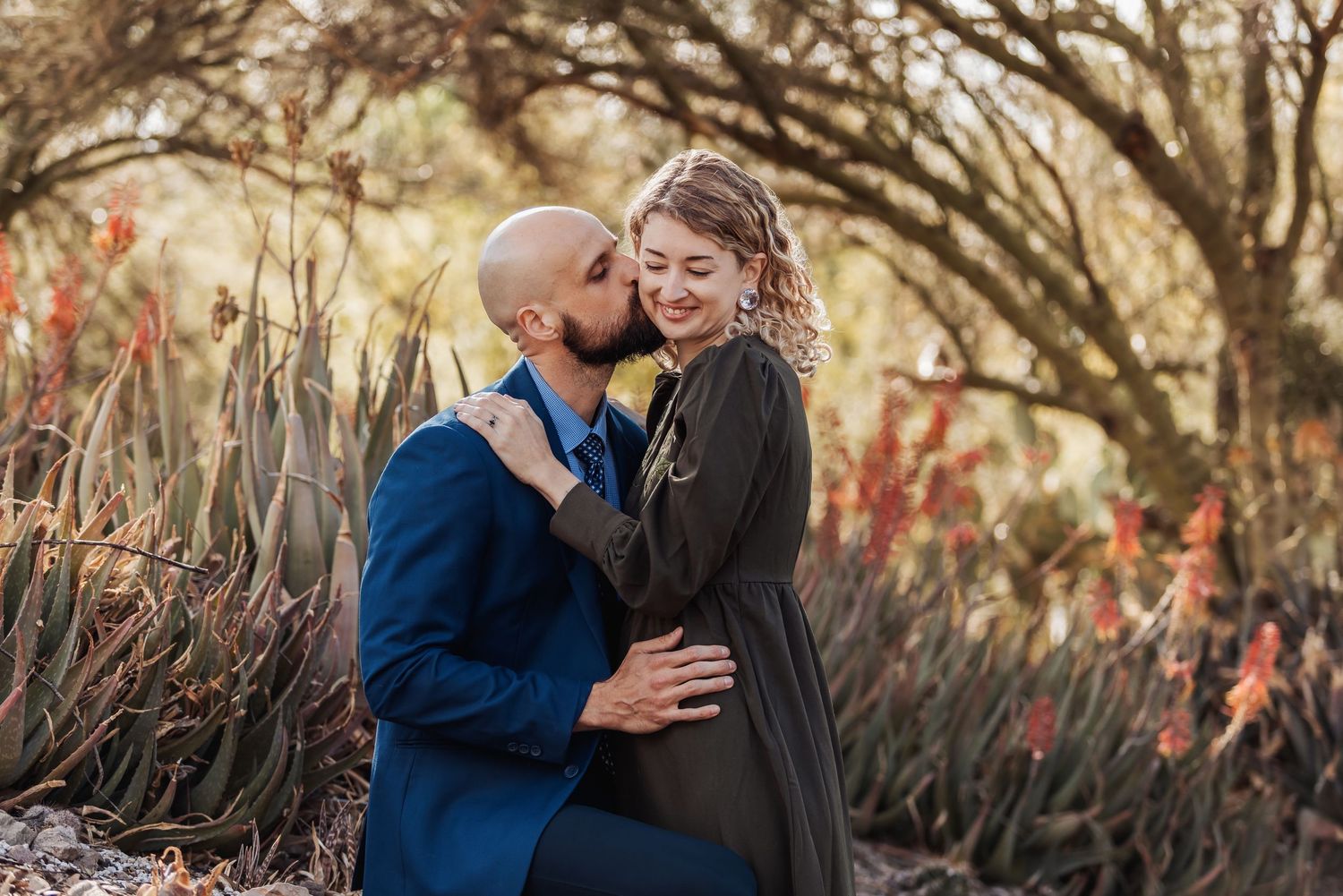A loving couple embraces in a garden setting with red aloe plants and natural bokeh lighting in the background.