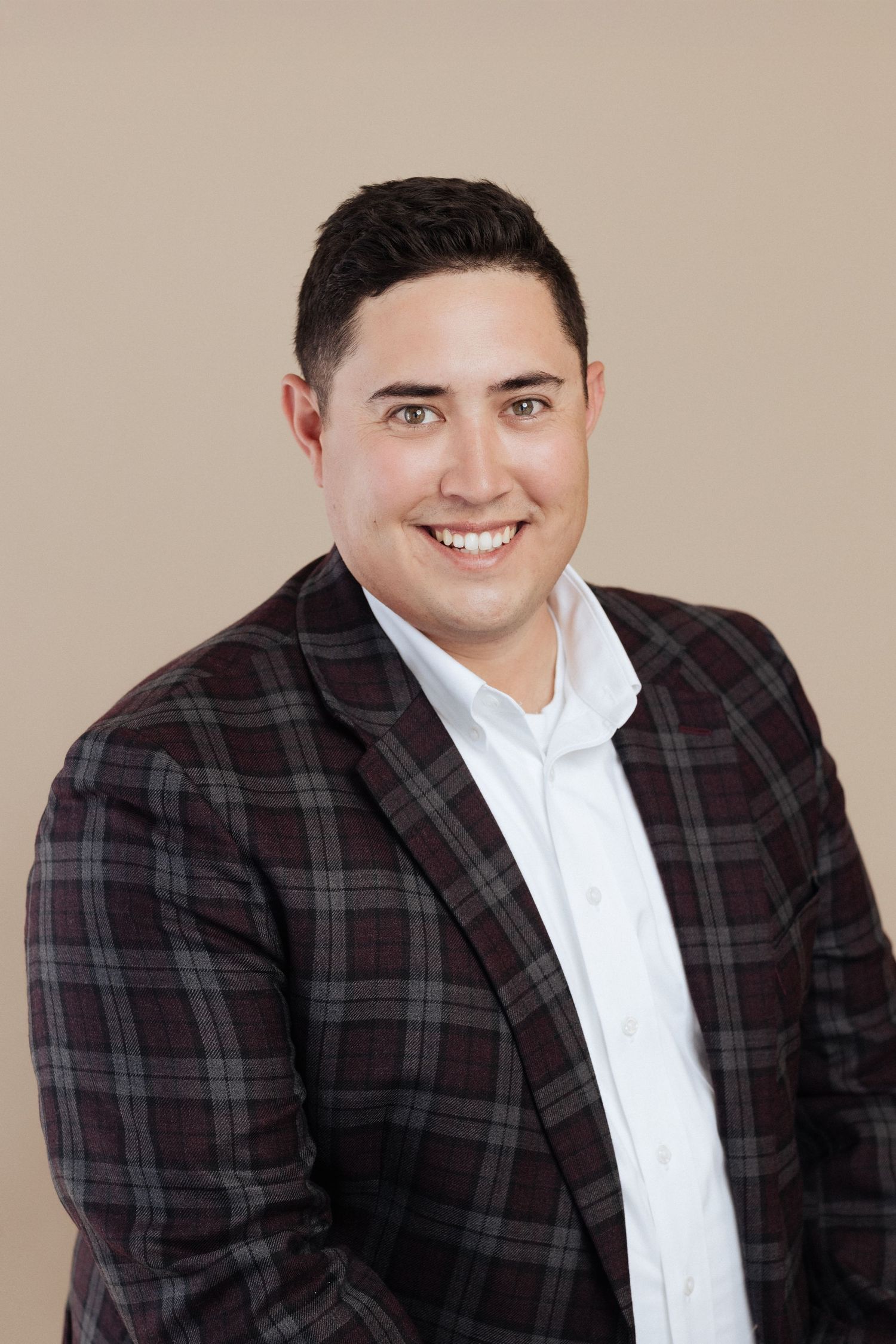 Professional headshot of someone wearing a plaid blazer and white dress shirt against a neutral beige background.