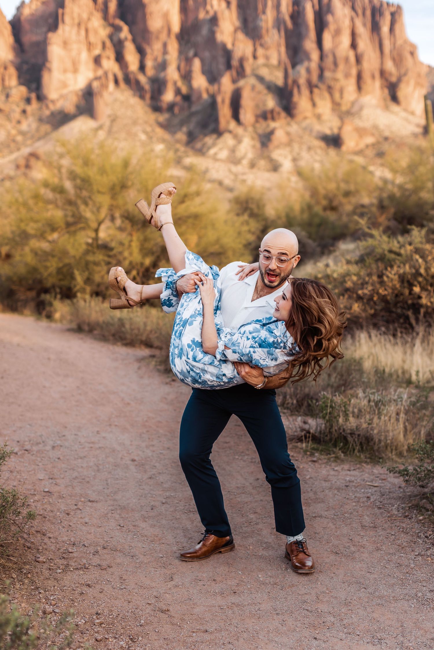 A playful couple shares a romantic moment on a desert trail with stunning mountain cliffs in the background.