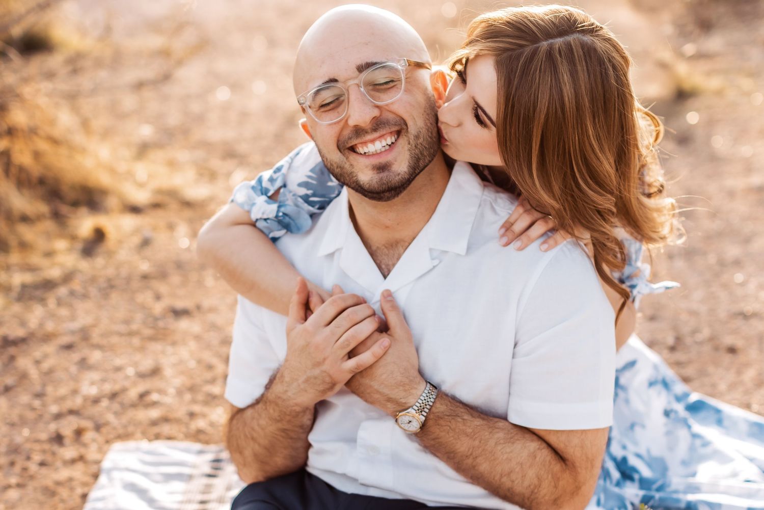 A romantic couple enjoys a tender moment together while sitting outdoors on a sunny day in a natural setting.