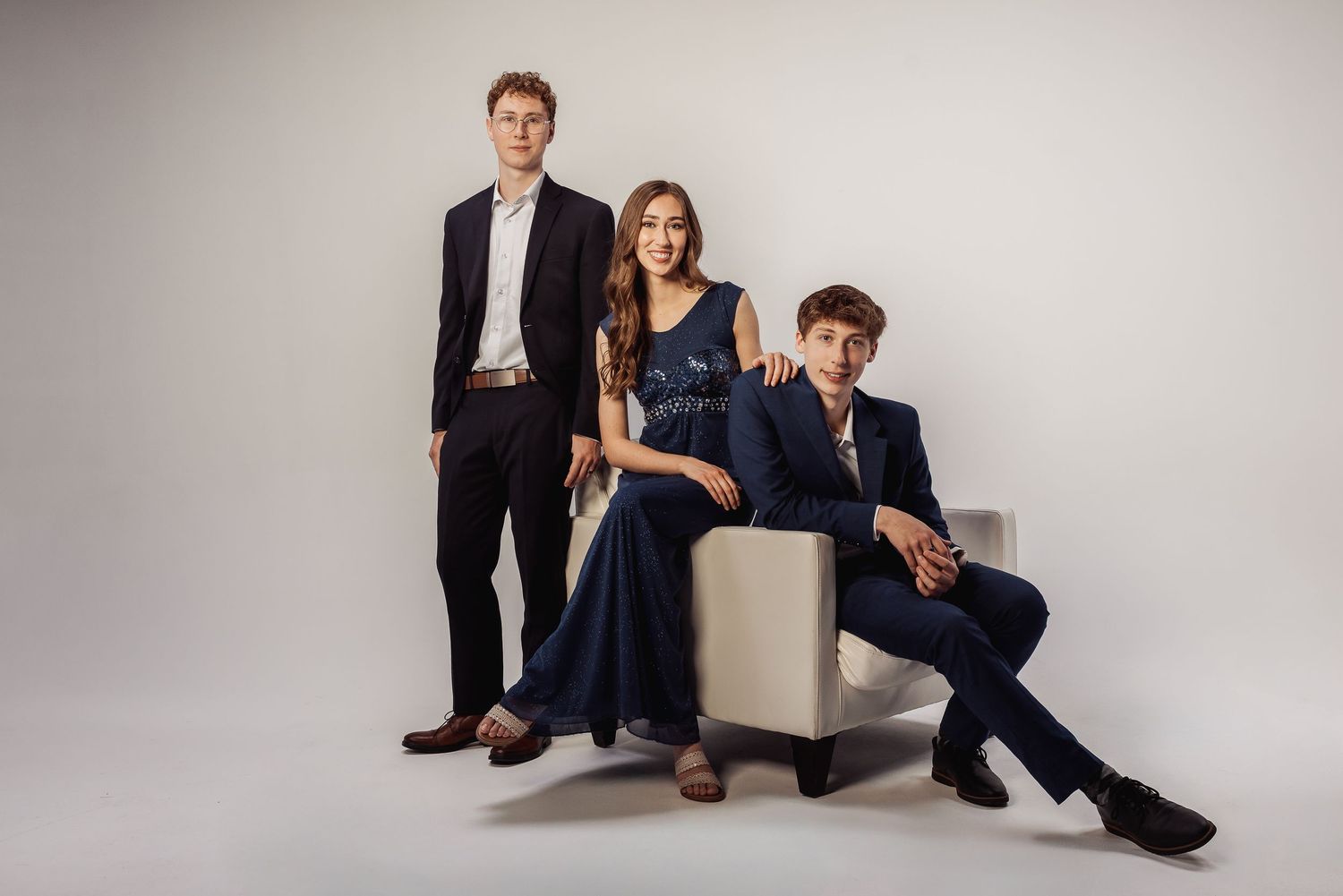 Three people in formal attire pose together on a light gray backdrop with one person seated on a white ottoman.