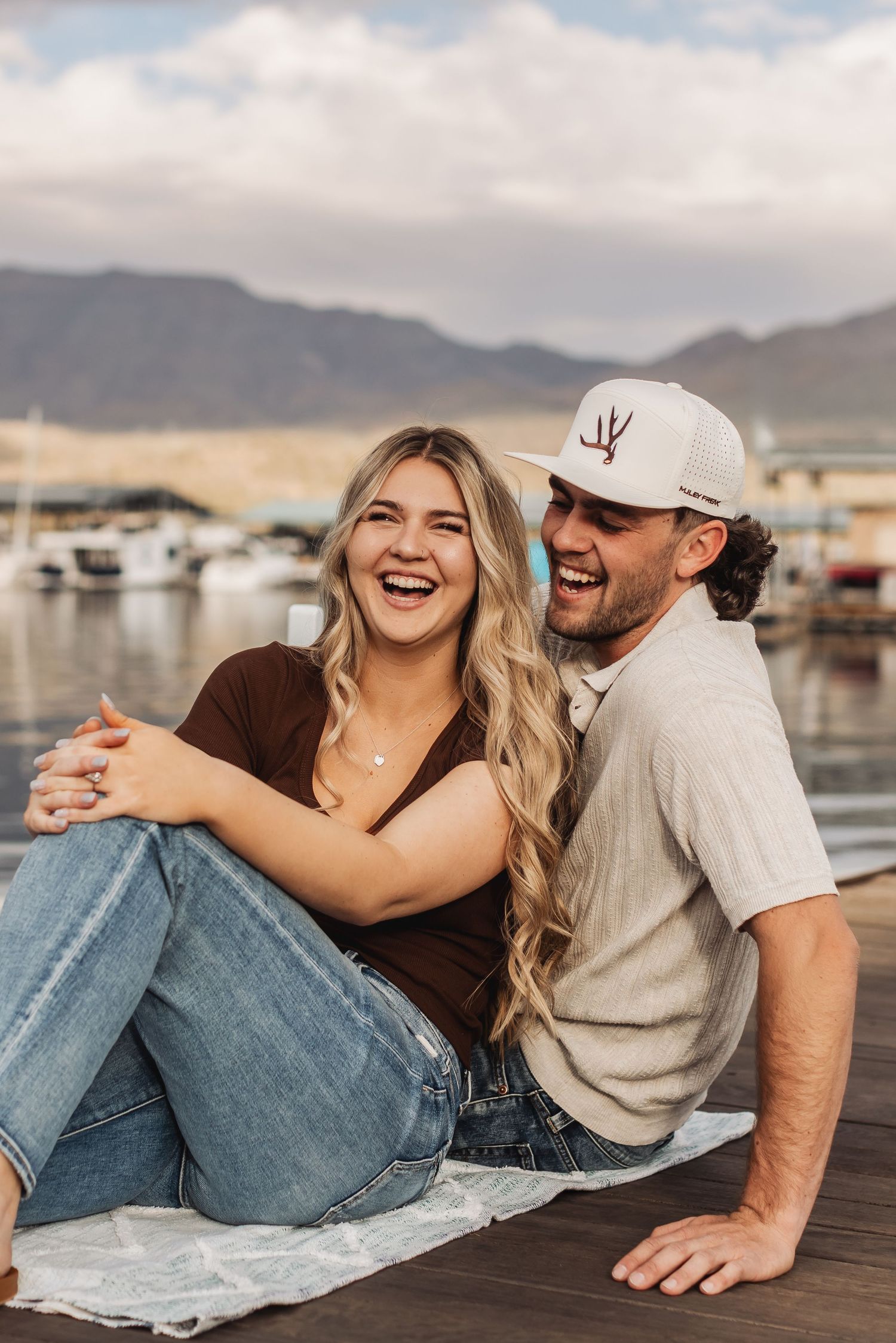 A joyful couple shares a candid laughing moment together while sitting on a dock at a marina with mountains in the background.