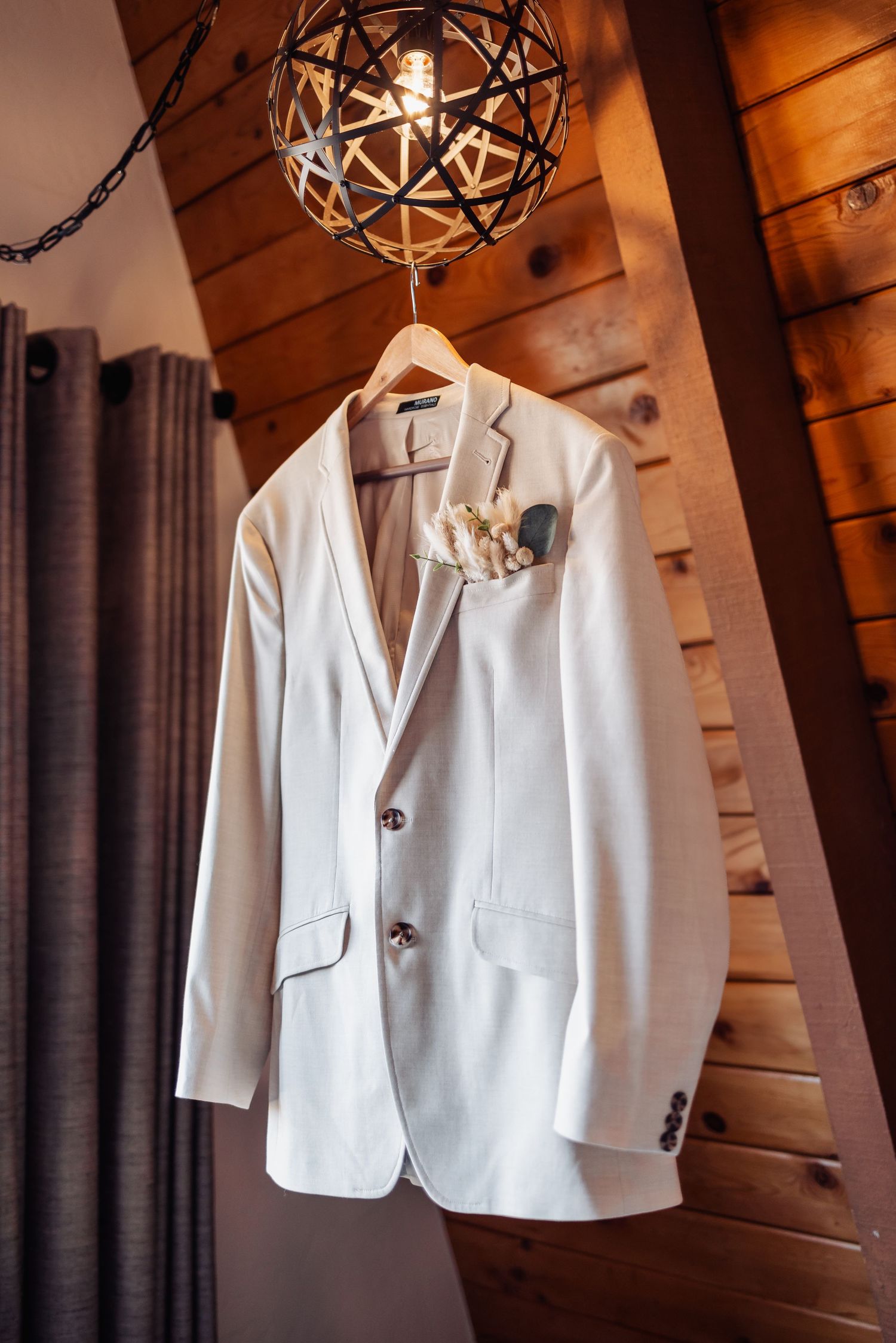 A white formal suit jacket hangs from a hanger beneath a decorative spherical light fixture against wooden walls.