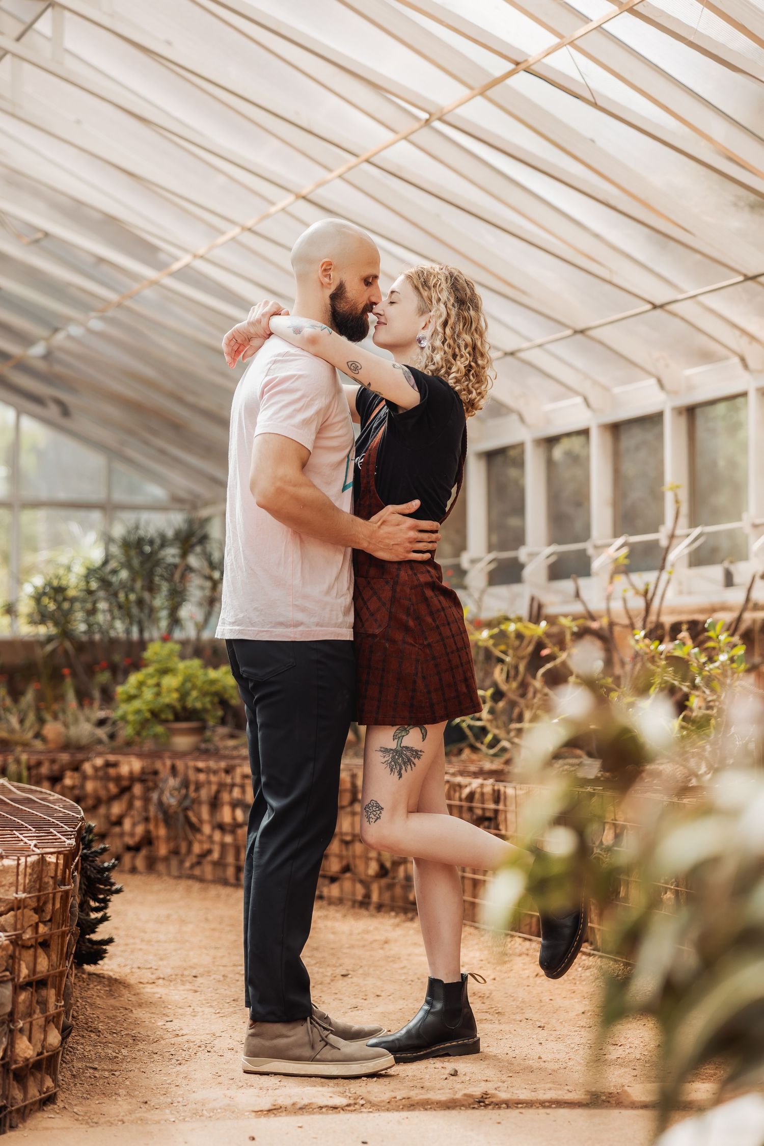 Romantic couple embraces in a sunlit greenhouse surrounded by plants and rustic decor.