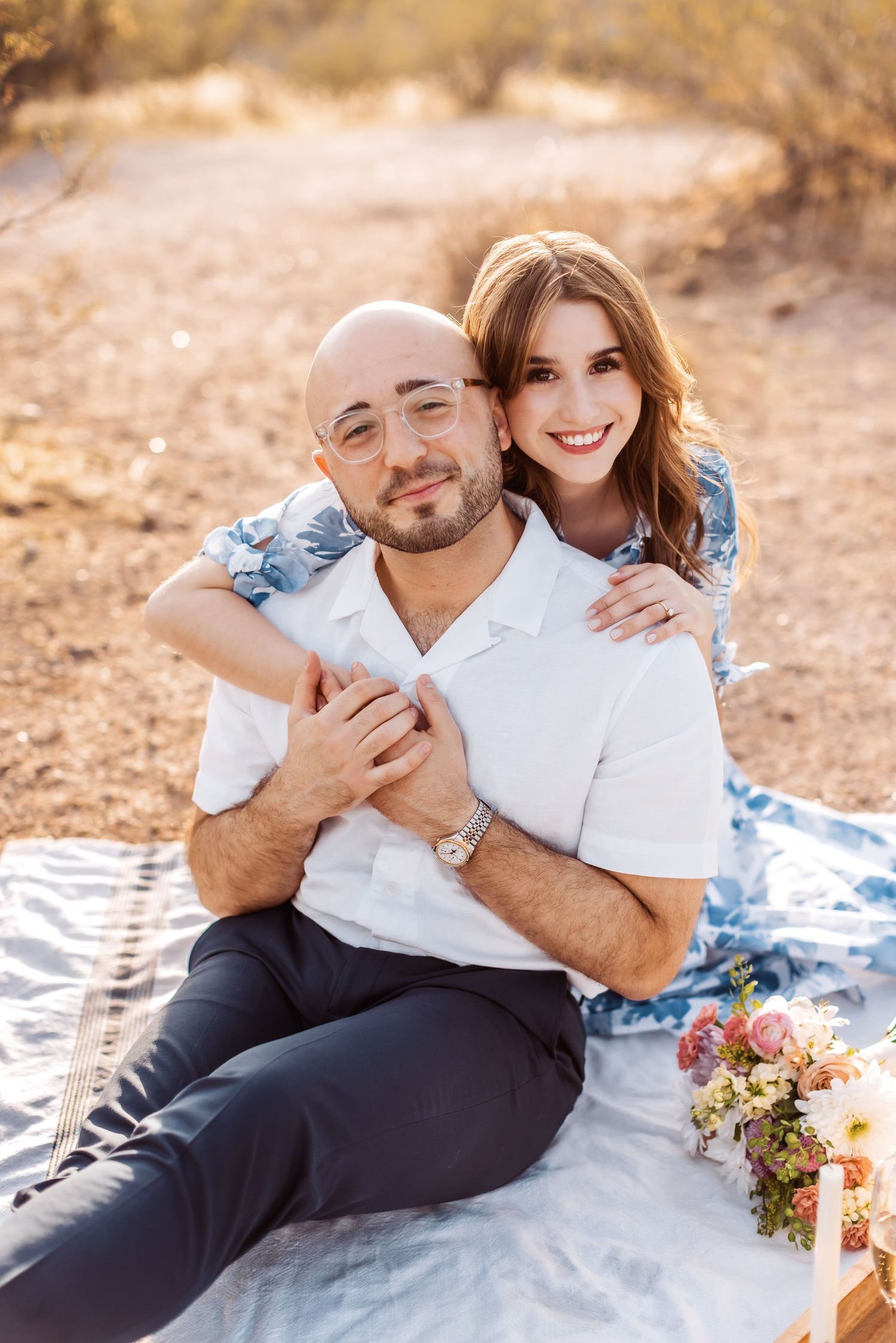 A couple sits together on a picnic blanket outdoors during a warm sunset photoshoot with flowers nearby.