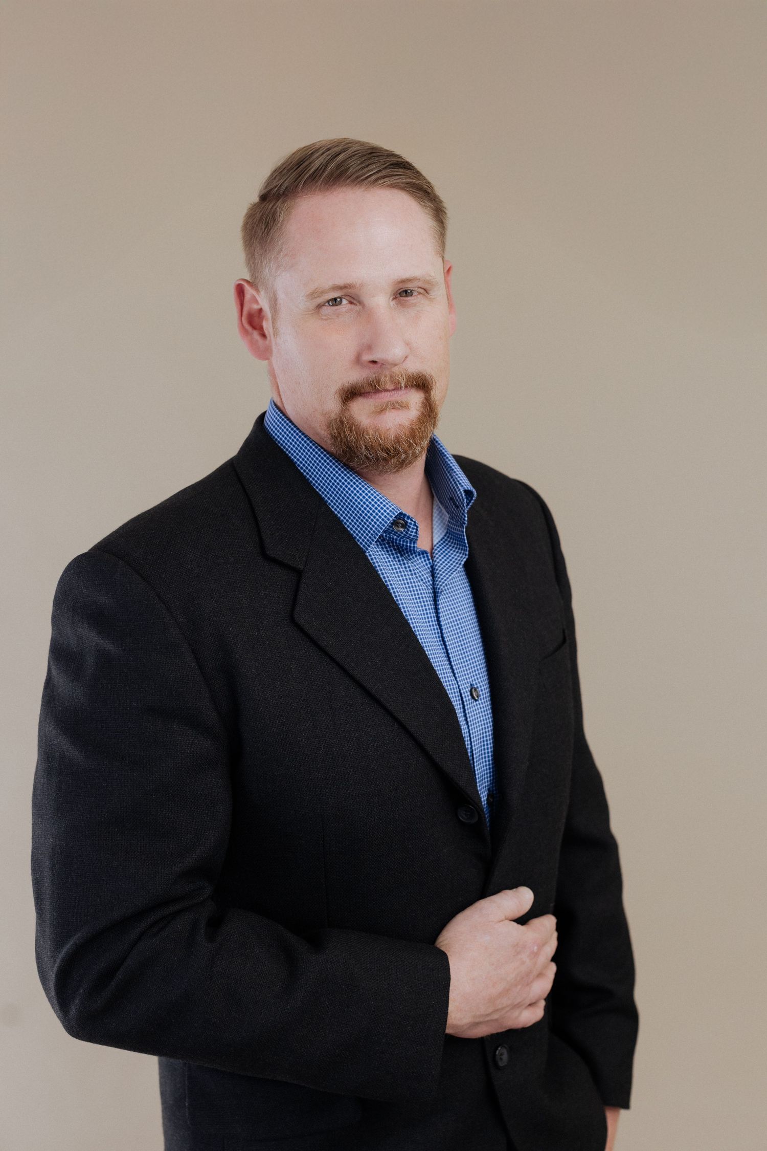 Professional headshot of person in black suit jacket and blue button-up shirt against neutral beige background.