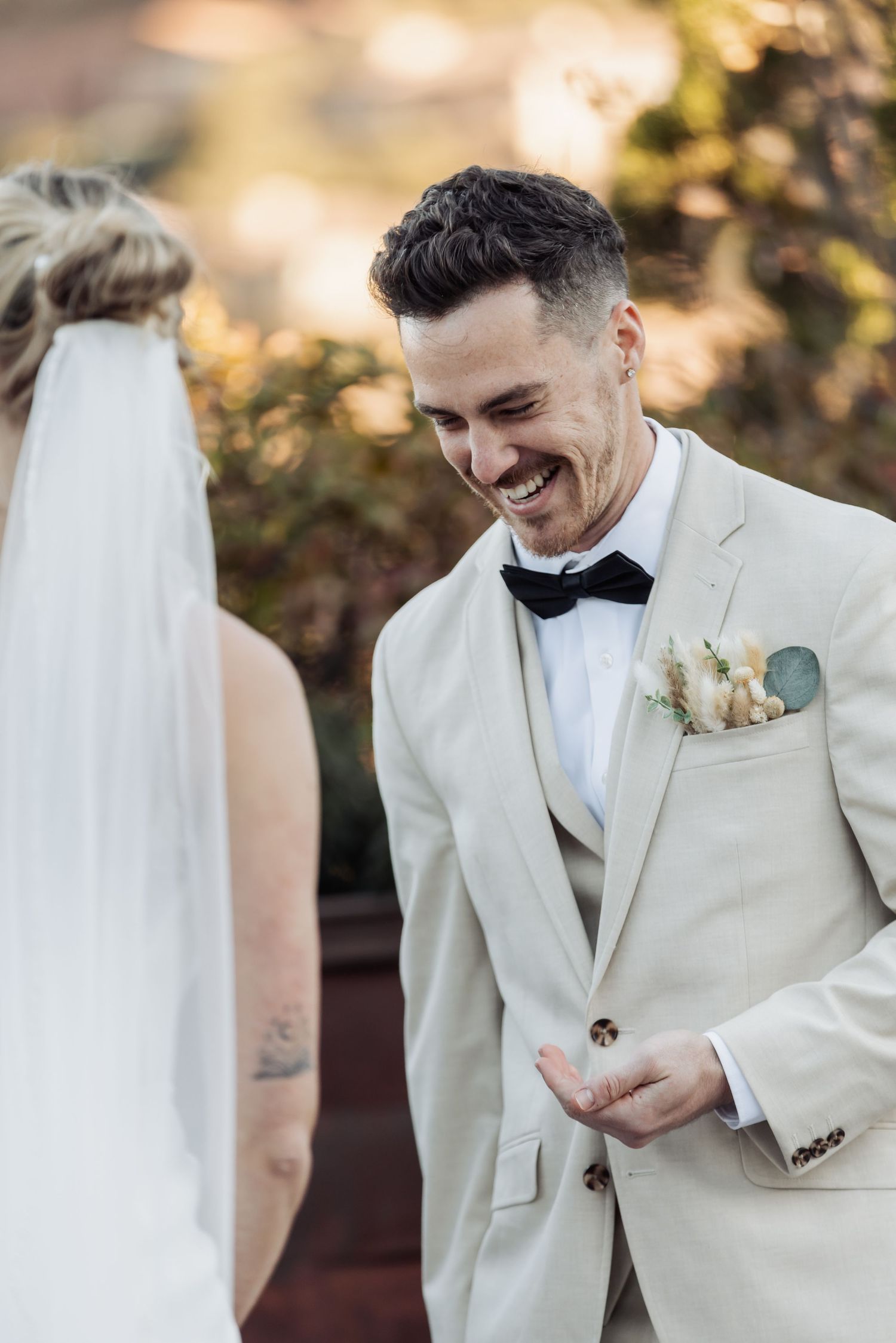 A groom in a cream suit and bow tie smiles joyfully during an outdoor wedding ceremony.