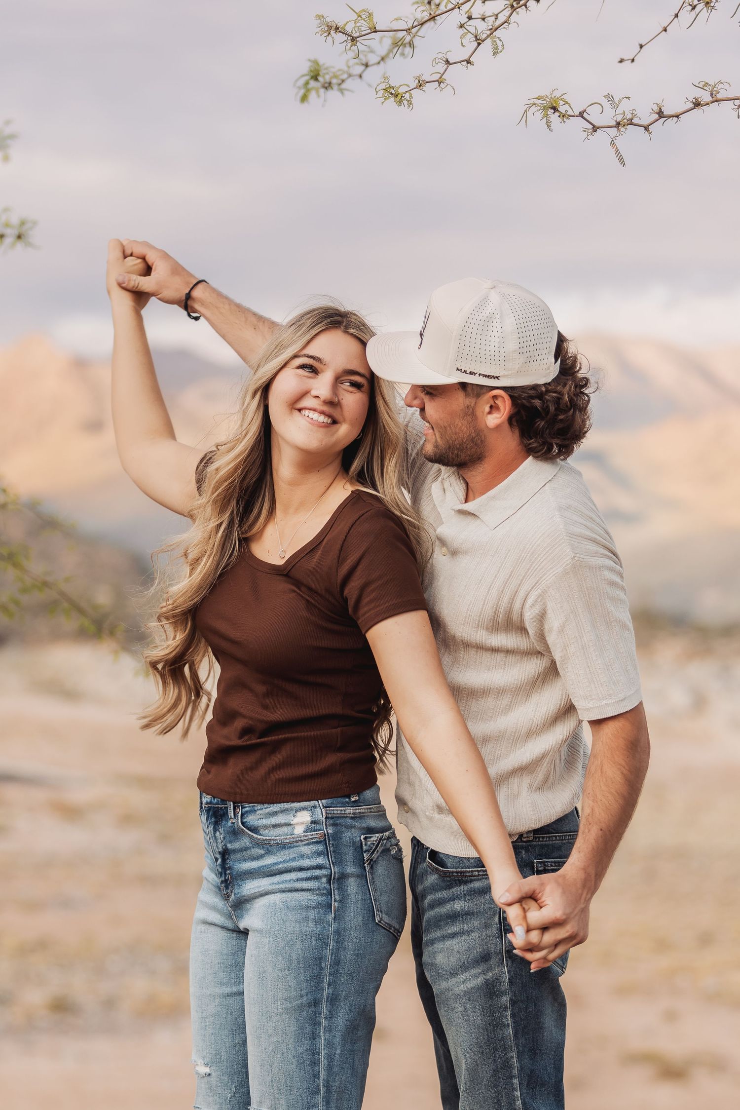 Couple dancing together in casual outfits against a desert mountain landscape during sunset.