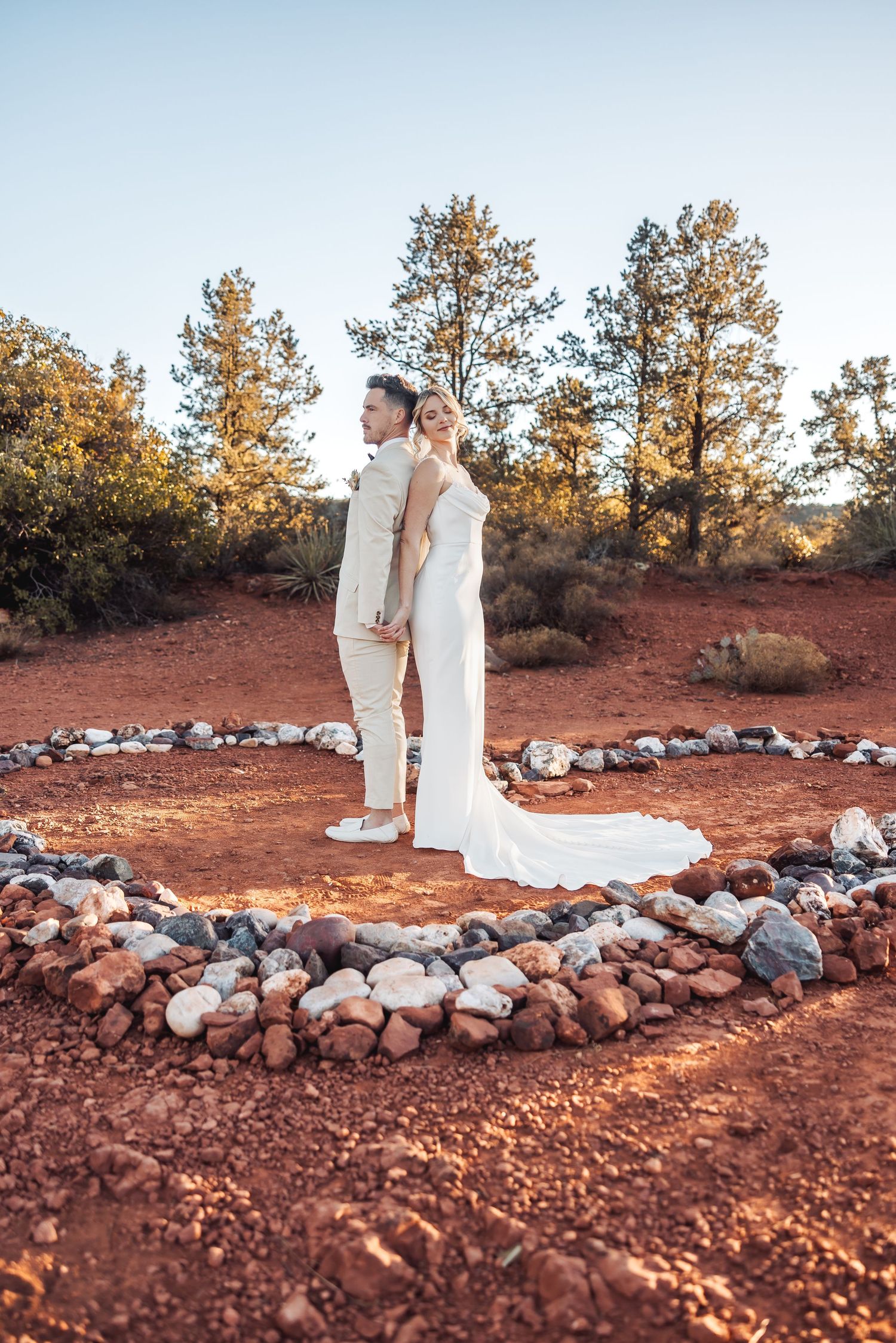 Couple in white attire poses back-to-back in a stone circle on red desert terrain at sunset.